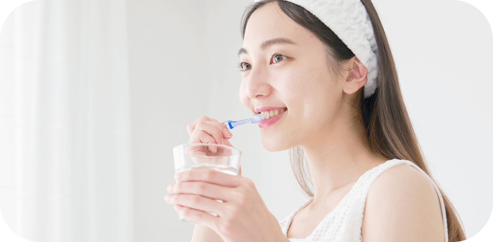 コップを手に持ちながら歯を磨く、ヘアバンドをつけた若い女性の笑顔